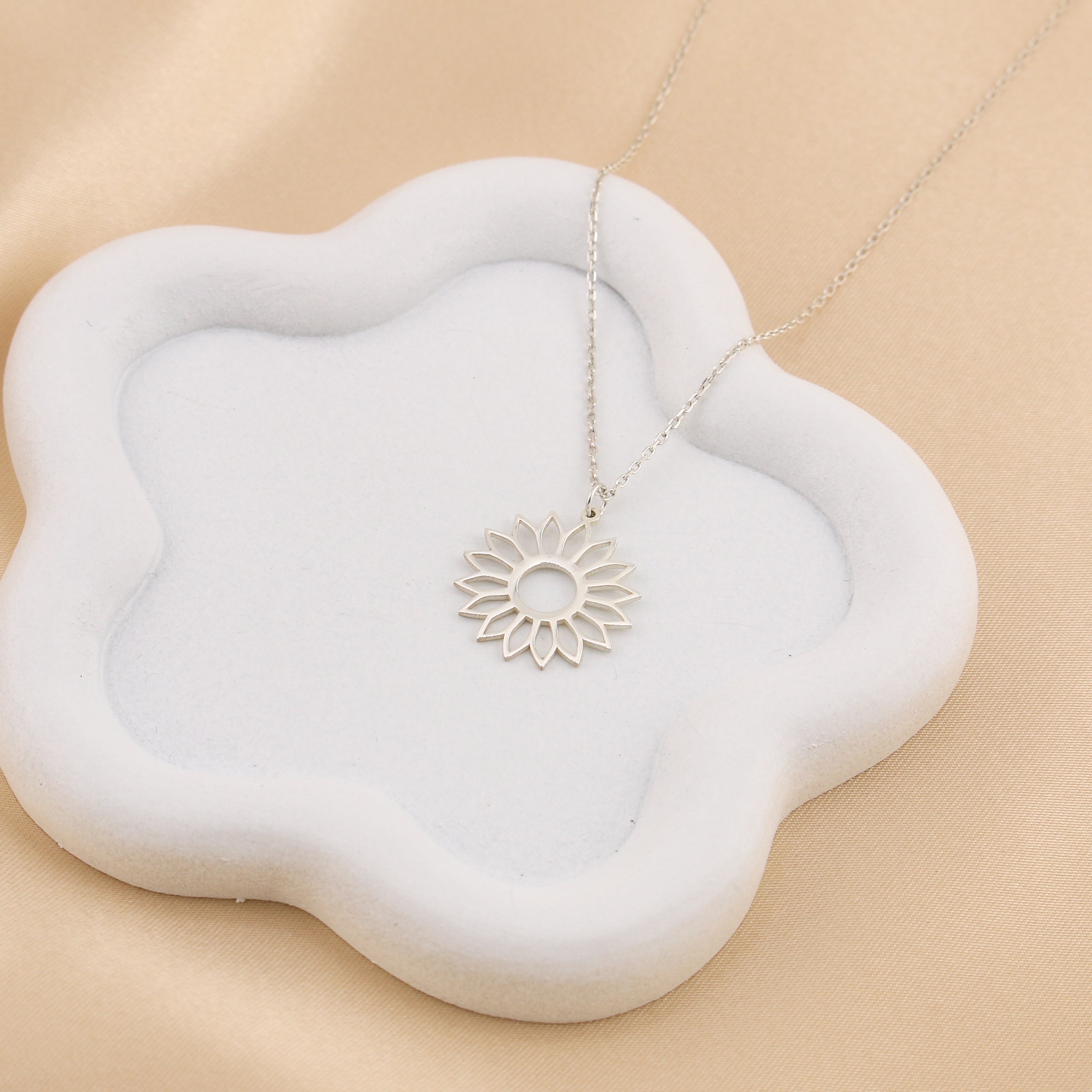 Silver necklace with a sunflower pendant on a white flower-shaped jewelry cushion against a beige fabric background.