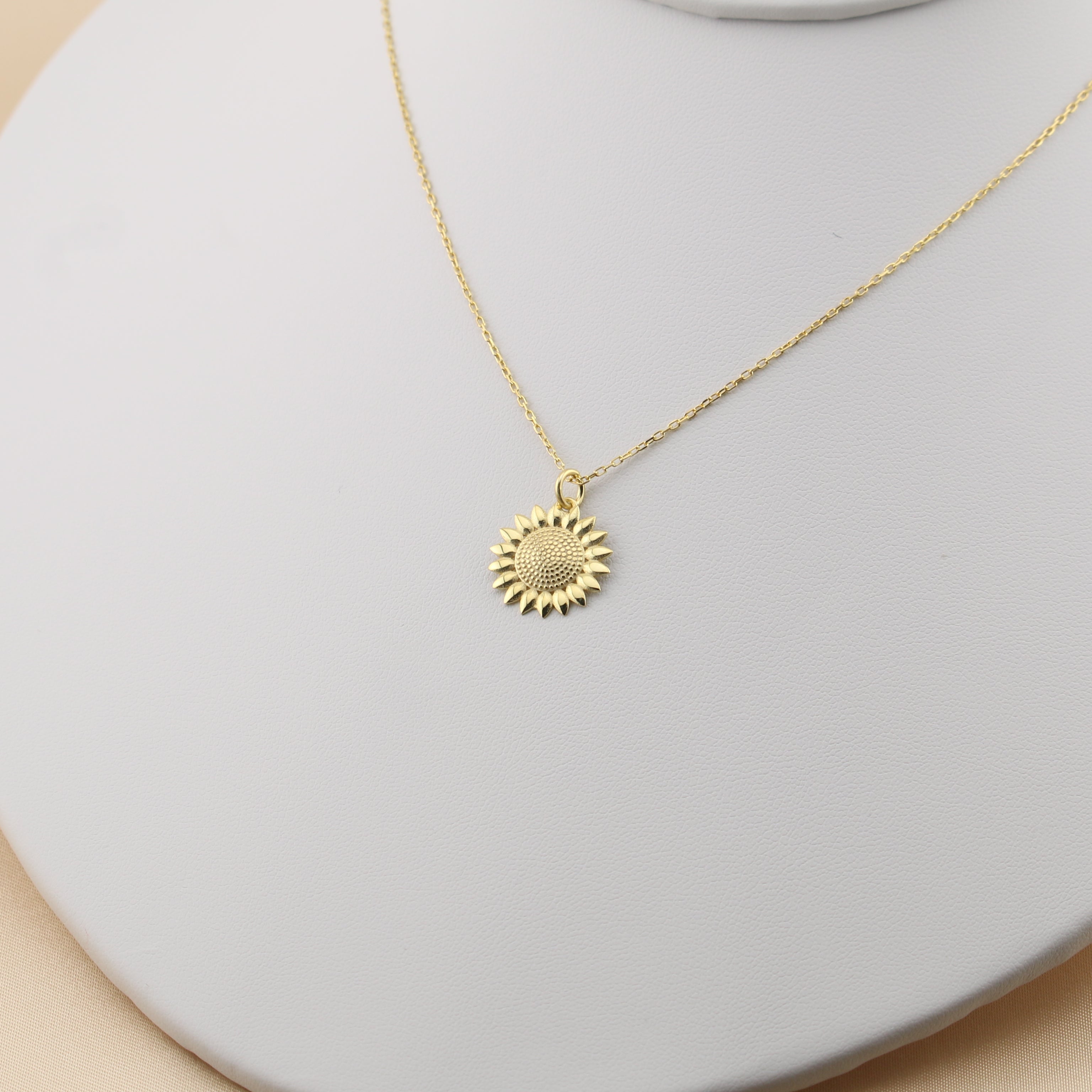 Gold necklace with a sunflower pendant on a white stand against a beige background